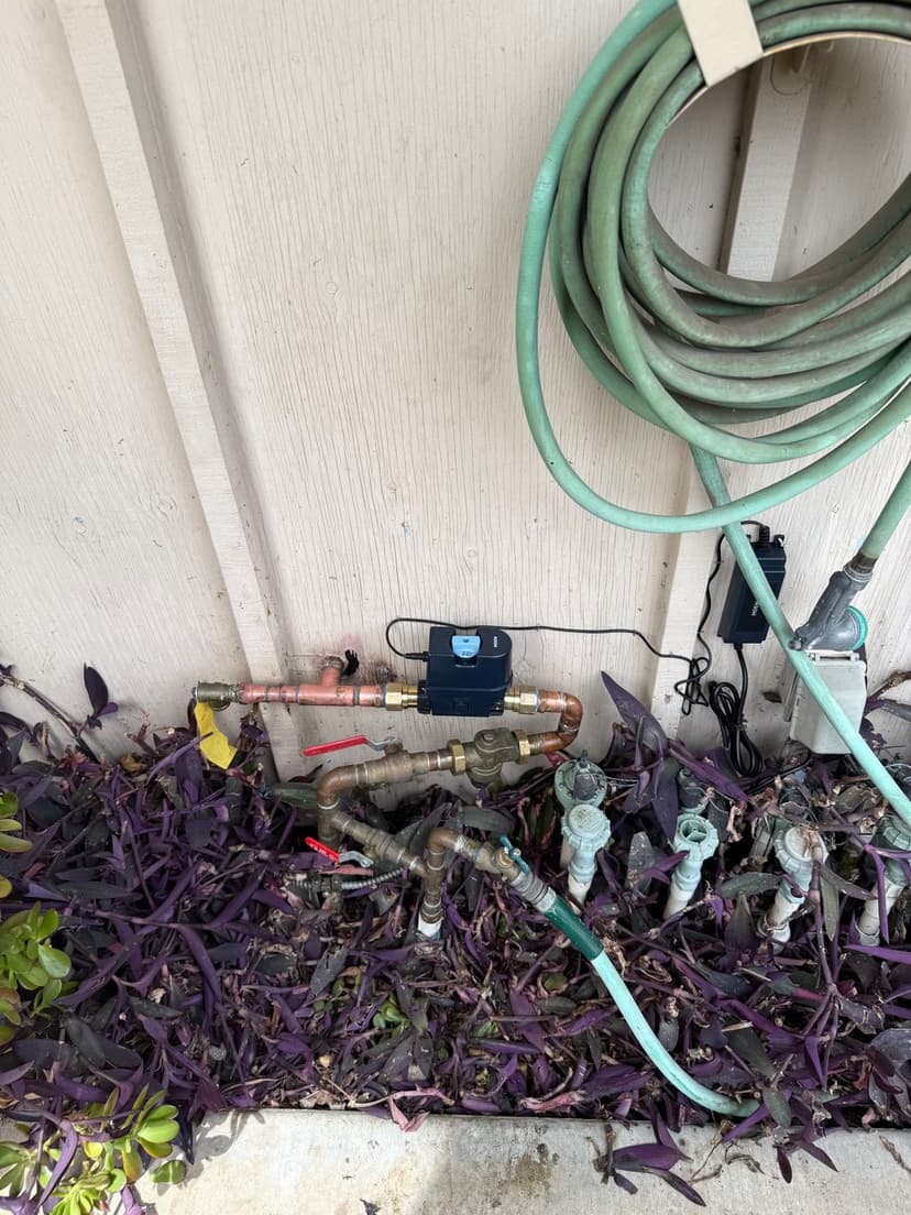 Outdoor plumbing setup with valves, hoses, and vegetation near a house wall.